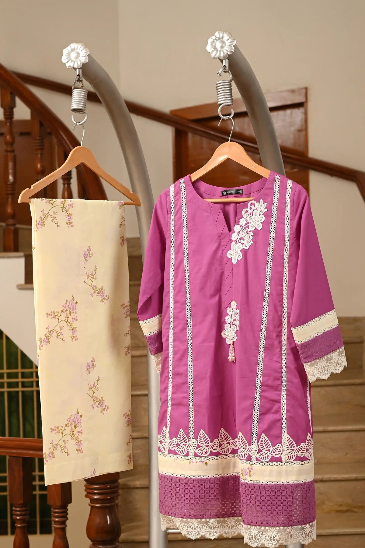 A purple traditional tunic with white lace embroidery hanging on a wooden hanger next to a cream floral skirt on a clothing rack in front of a wooden staircase.
